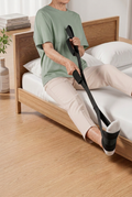 Older woman in a sage-green T-shirt using a black leg lifter strap while seated on a bed beside light-oak flooring